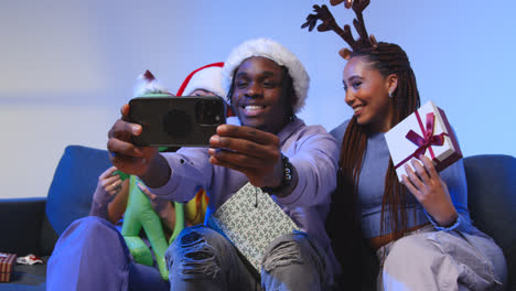 Studioaufnahme-Von-Freunden-Der-Generation-Z-Zu-Weihnachten,-Die-Mit-Weihnachtsmütze-Und-Rentiergeweih-Auf-Dem-Sofa-Sitzen-Und-Ein-Selfie-Mit-Dem-Mobiltelefon-Machen-1
